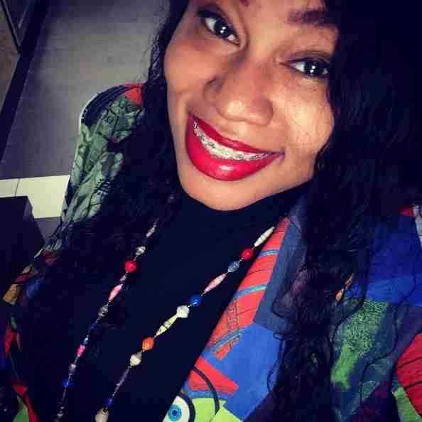 A person with long curly hair and braces smiles, wearing a colorful jacket and a beaded necklace.