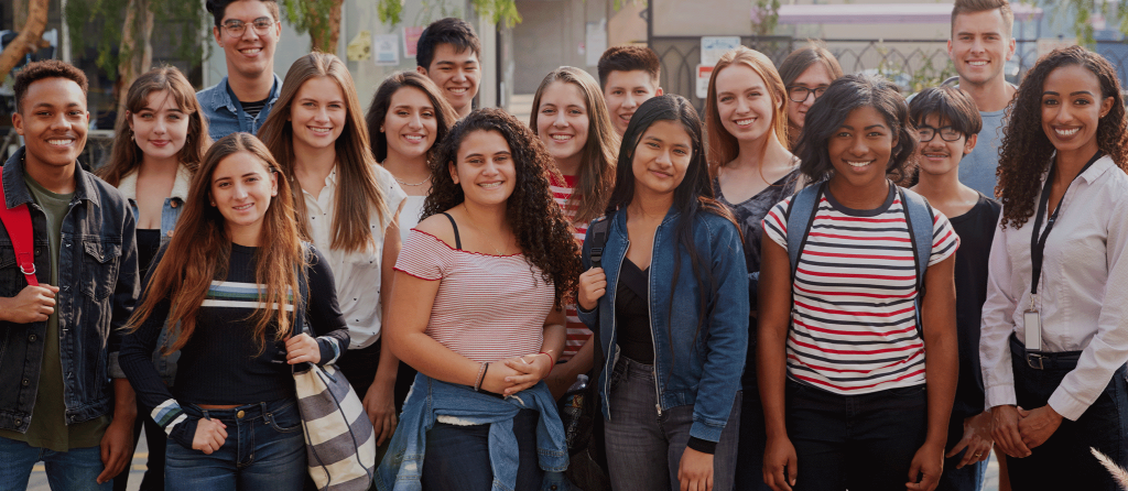 A group of young adults and teenagers standing outdoors, smiling at the camera. They appear to be in a casual setting, with trees and a fence in the background.