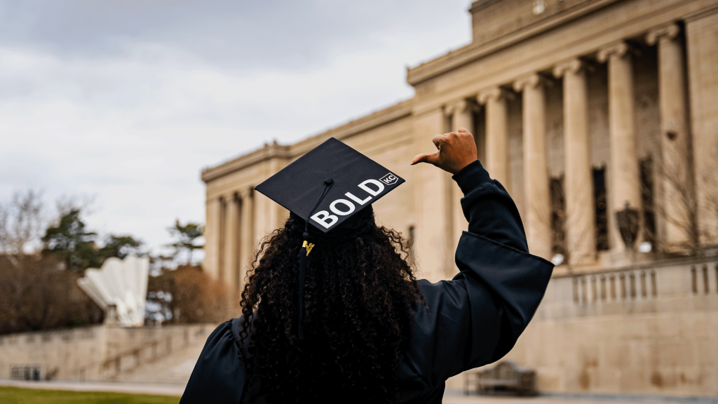 Person wearing a graduation cap with the word "BOLD" on it, raising their hand outside a classical-style building.