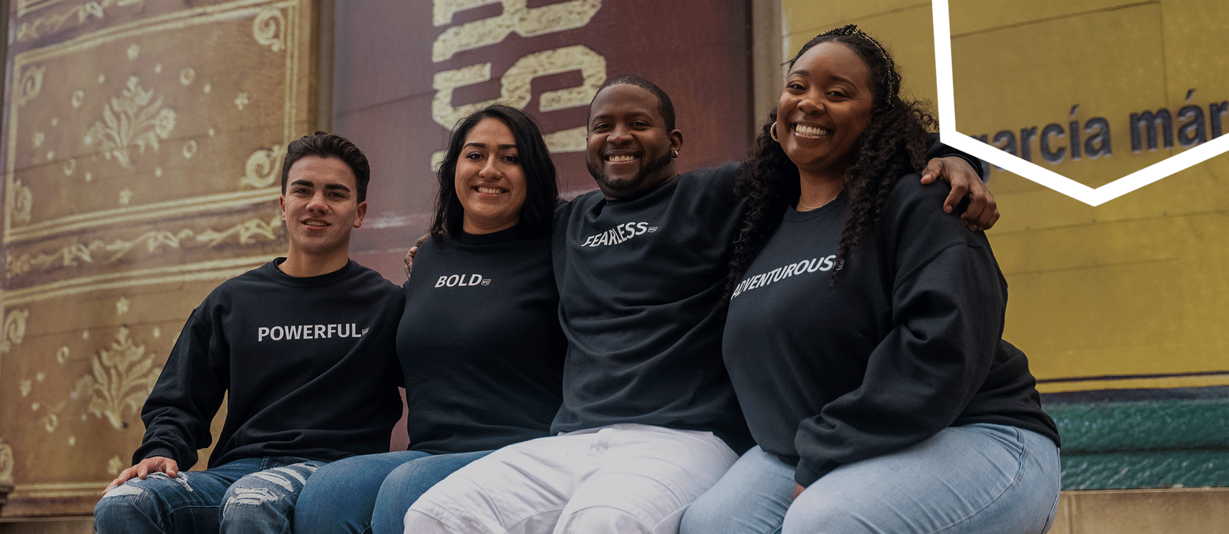 Four individuals are sitting on a ledge, smiling. They are wearing matching black sweatshirts with the words "POWERFUL," "BOLD," "FEARLESS," and "COURAGEOUS" printed on them.