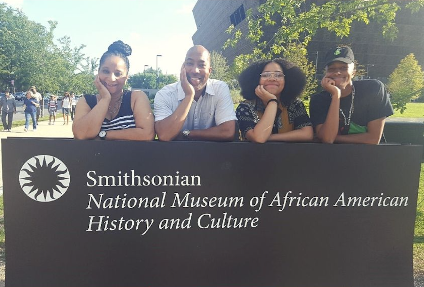 Four people pose behind a sign for the Smithsonian National Museum of African American History and Culture on a sunny day.