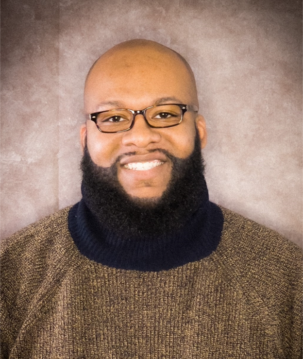 A man with a beard, glasses, and wearing a brown sweater smiles in front of a brownish-gray background.