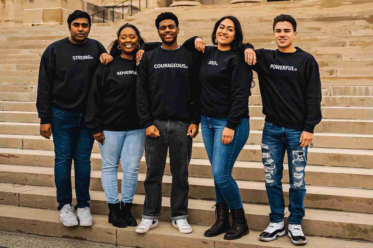 Five people stand side by side on steps, each wearing a black sweatshirt with words: "STRONG," "ADVENTUROUS," "COURAGEOUS," "KIND," and "POWERFUL." They are smiling with arms around each other.