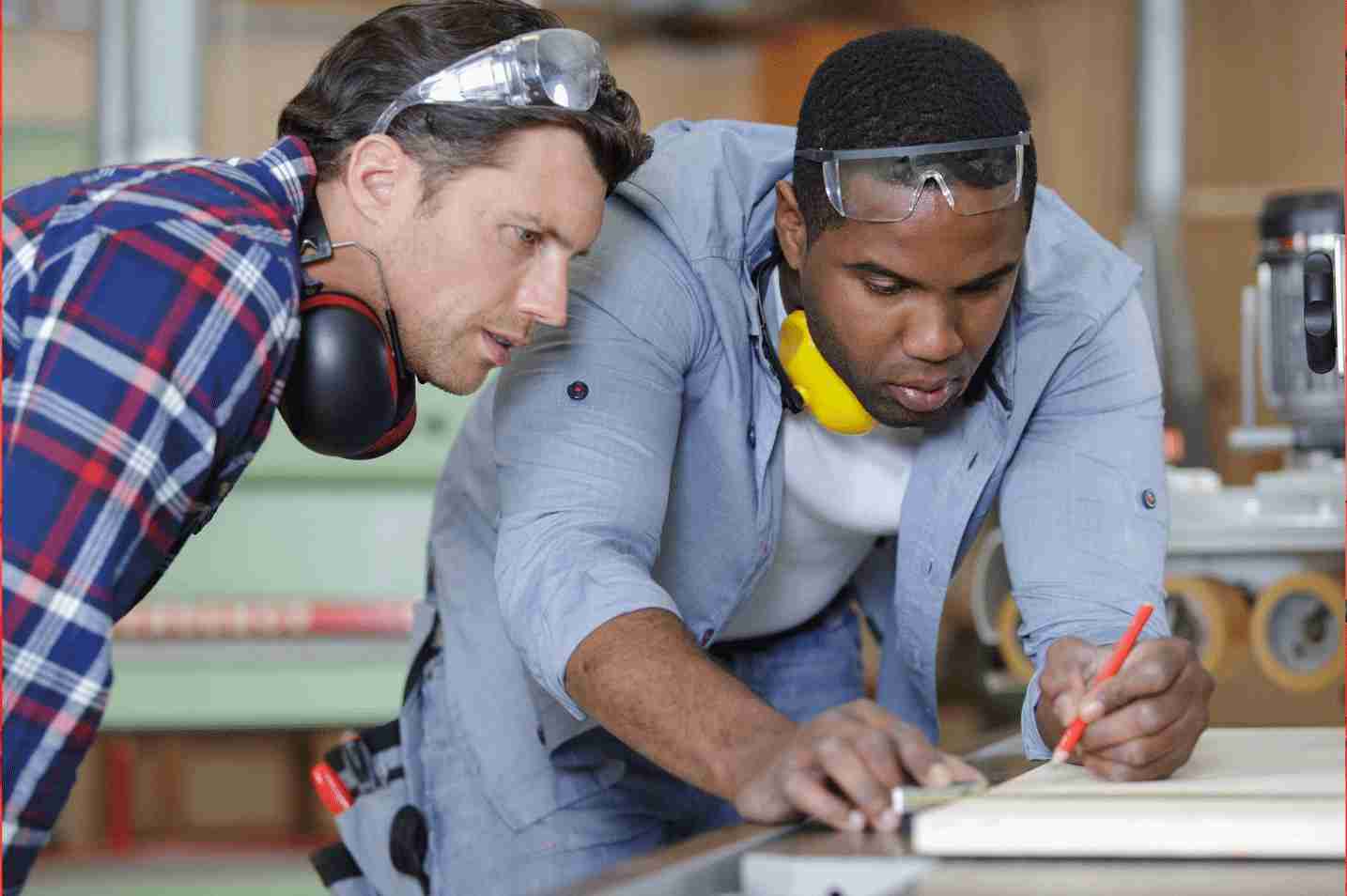Two men in a workshop concentrating on measuring and marking a wooden board, wearing protective eyewear and ear protection.