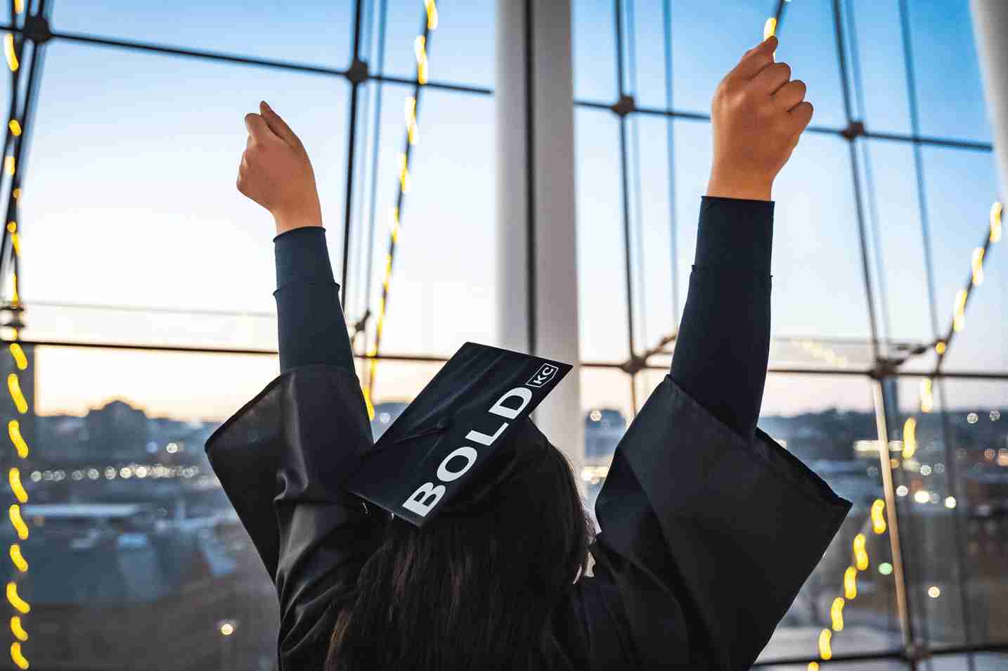 A person wearing a graduation cap with the word "BOLD" on it raises their arms in celebration in front of large windows with a cityscape in the background.
