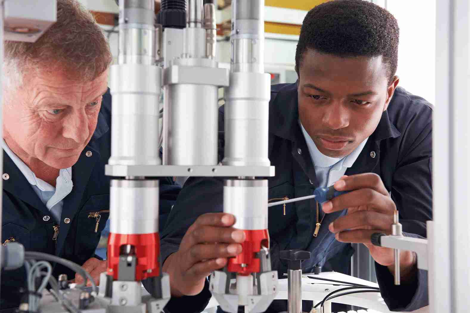 Two engineers work together on a piece of industrial machinery, focusing on adjustments and components, in a well-lit workshop.