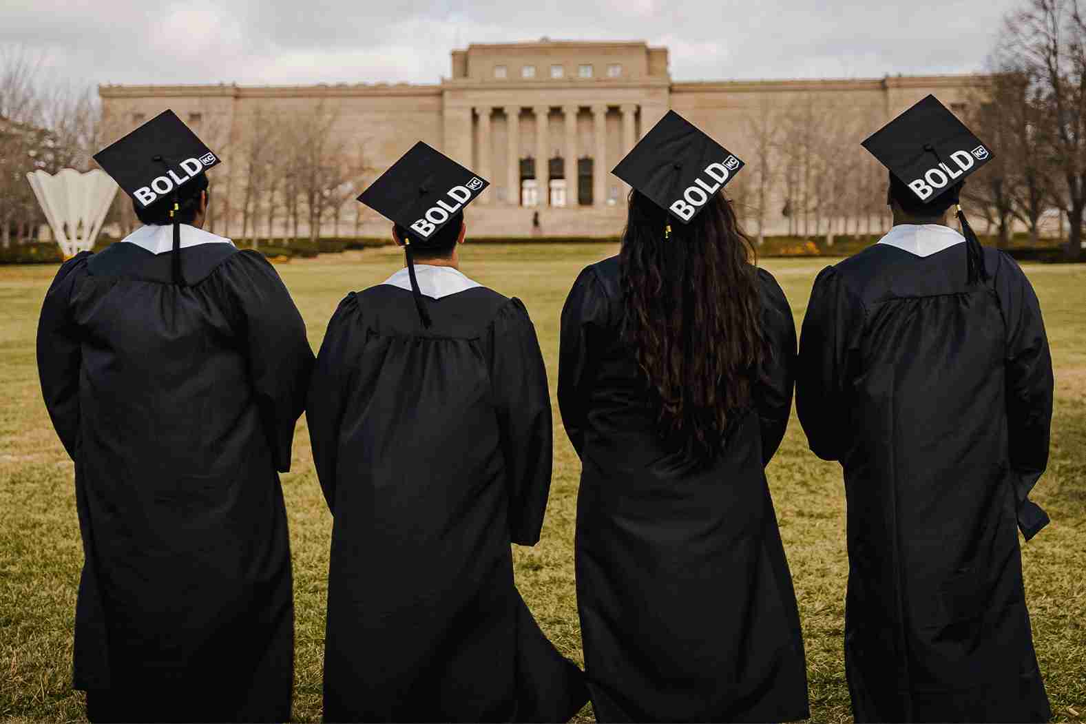 Four graduates in black gowns and caps with "BOLD" text on them stand with their backs facing a large building.