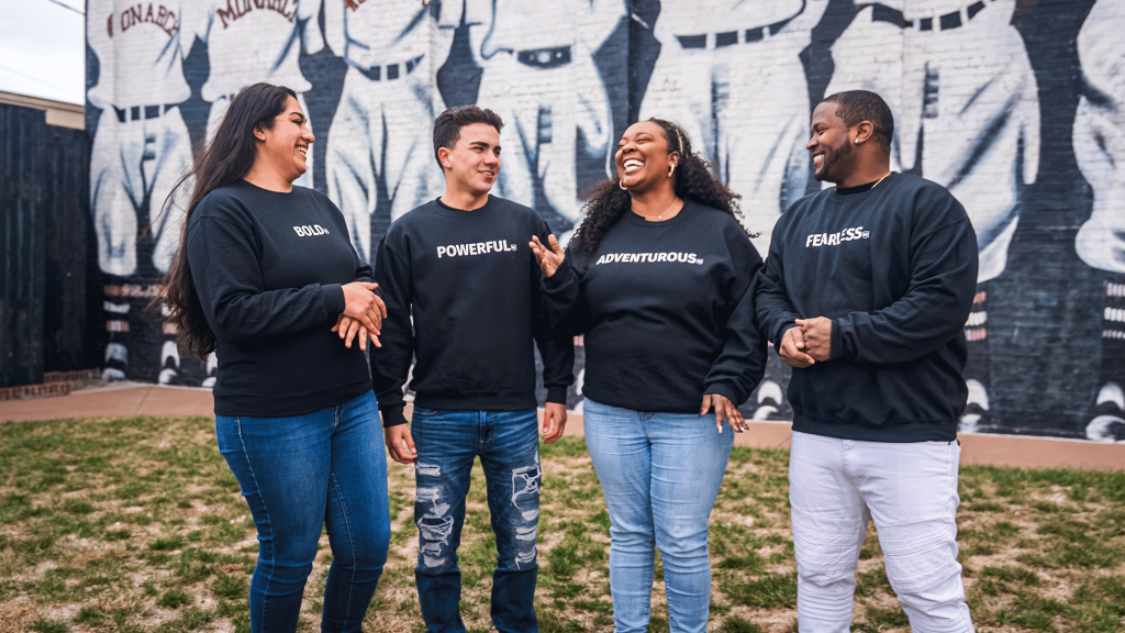 Four people stand outdoors in front of a mural, wearing black sweatshirts with different words on them: "Bold," "Powerful," "Adventurous," and "Fearless." They are smiling and engaged in conversation.