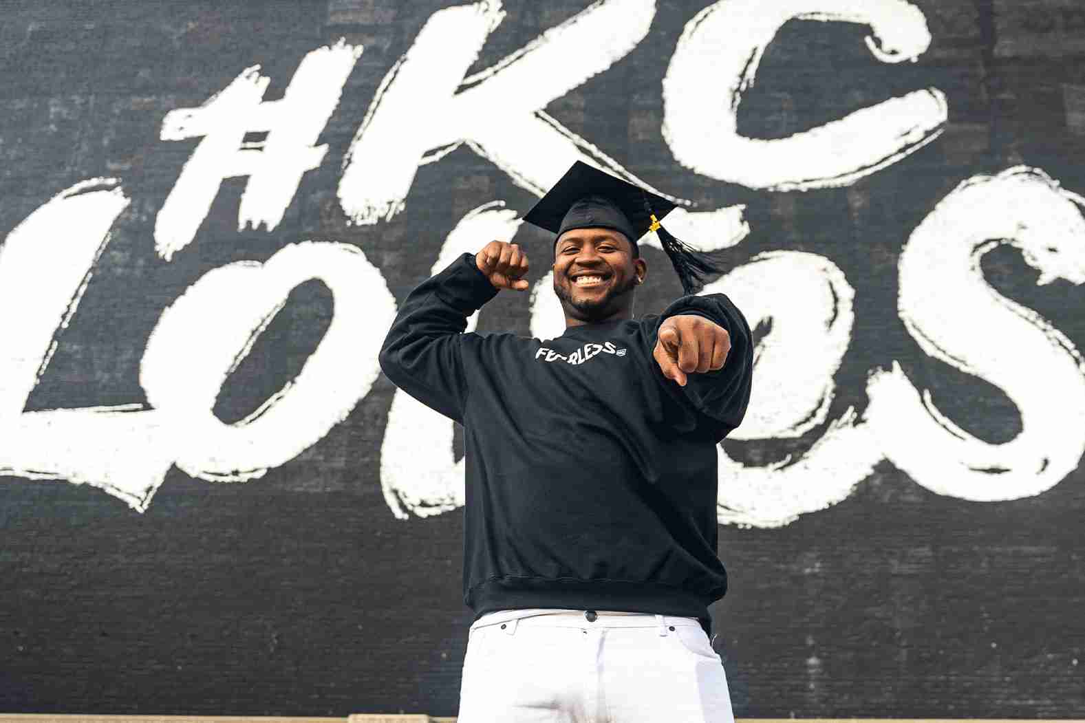 A smiling individual wearing a graduation cap points towards the camera, standing in front of a large mural with "#KC Loves" painted on it.