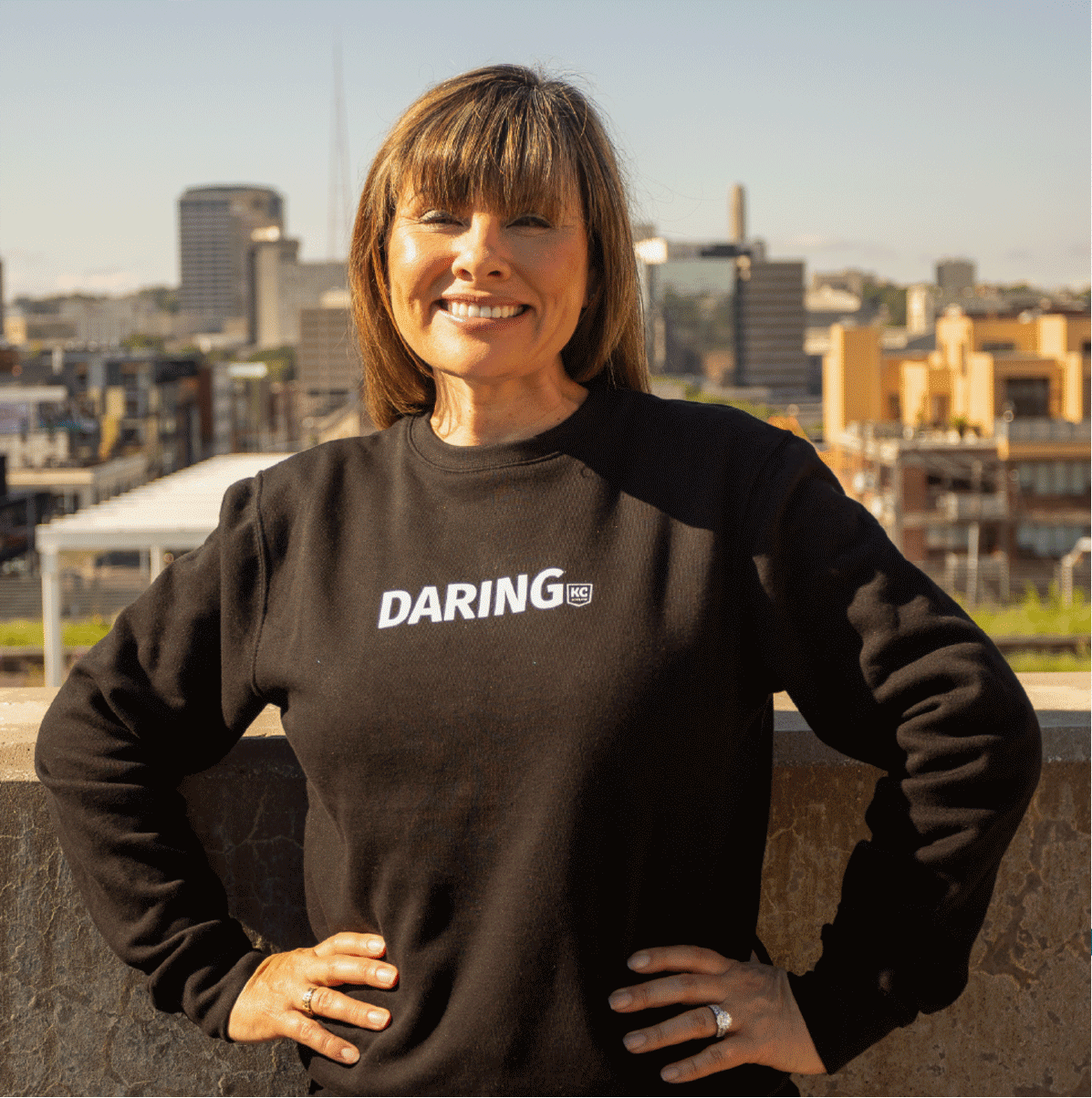 A woman wearing a black sweatshirt with the word "DARING" stands outside against a backdrop of city buildings, smiling with her hands on her hips.