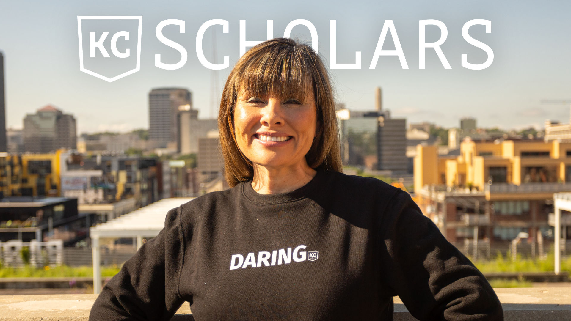 A person stands outdoors smiling with a cityscape in the background. They are wearing a black sweatshirt with the word "DARING" and the KC Scholars logo is visible above them.