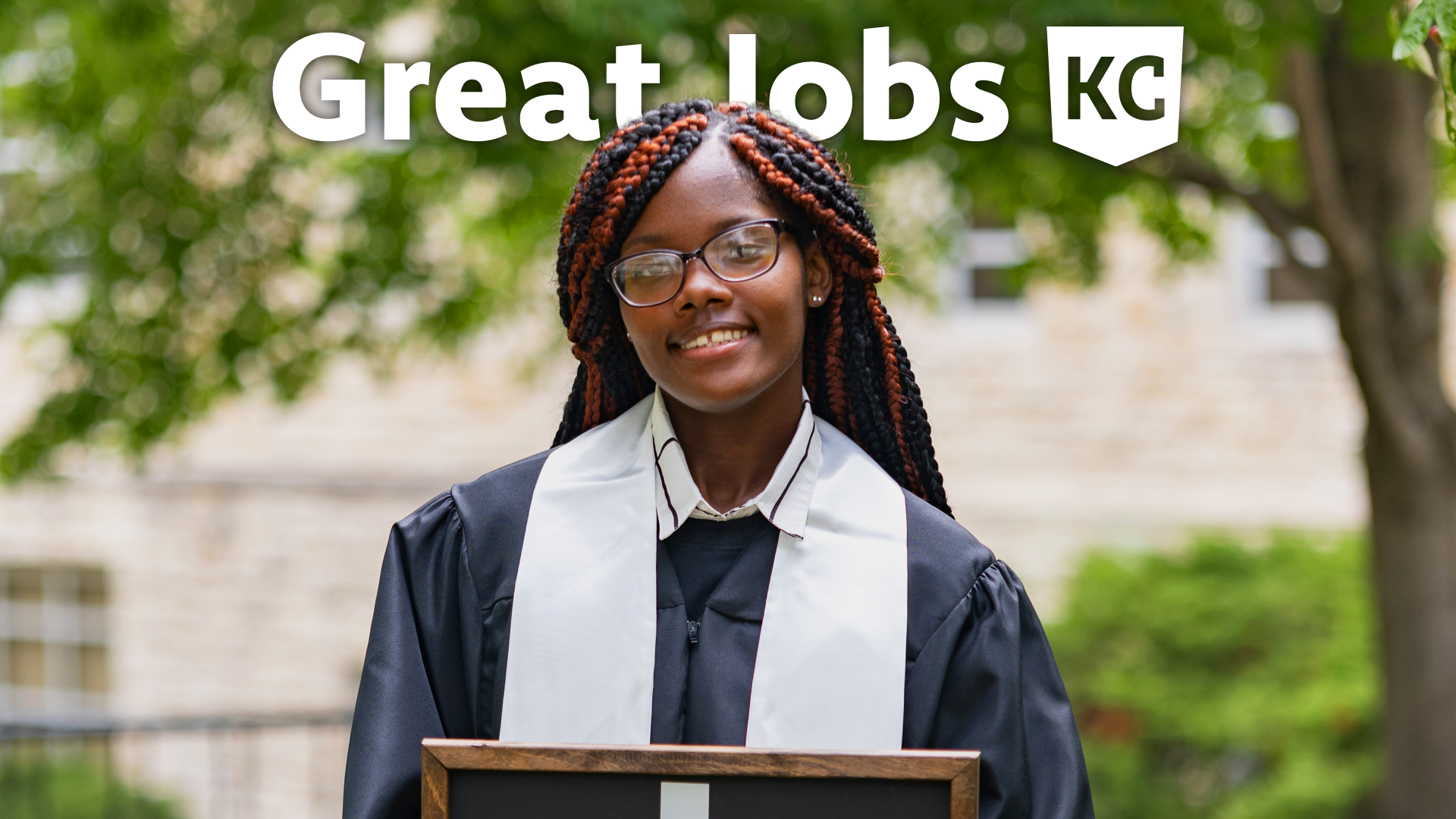 A person wearing graduation attire and holding a certificate stands outdoors in front of a tree. Above their head, the text reads "Great Jobs KC," celebrating new careers.