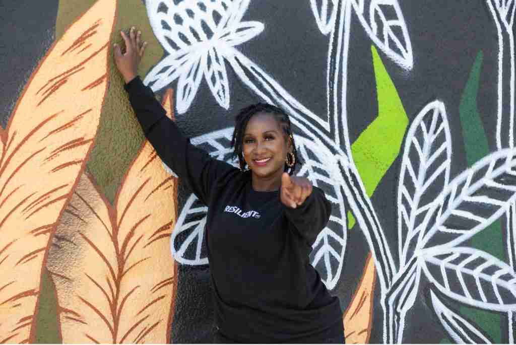 A person in a black sweatshirt stands in front of a colorful mural with plant designs, one hand on the wall and the other pointing forward, smiling confidently as if ready to take on new jobs.