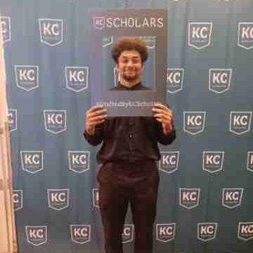 A person holds a large frame that reads "#KCScholars" at a backdrop with the KC Scholars logos, celebrating their scholarship achievement and future career aspirations.