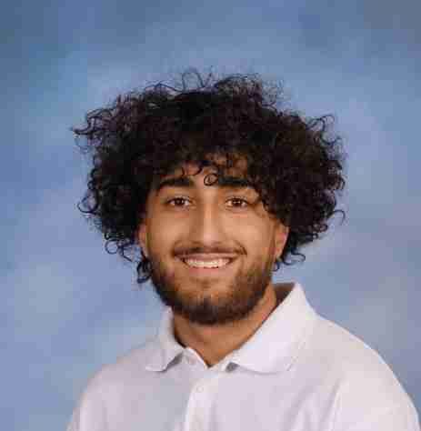 A young man with curly hair and a beard smiles at the camera against a blurred blue background. He is wearing a white collared shirt, perhaps excited about new career opportunities or having recently secured a scholarship.