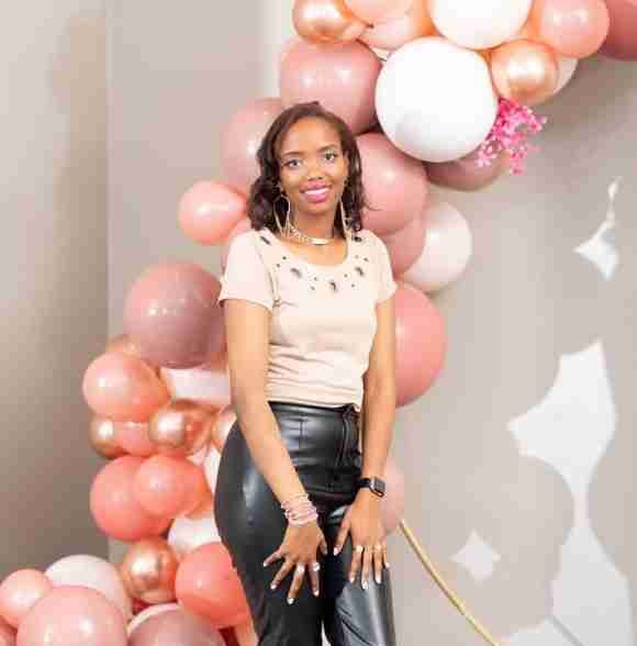 A woman stands in front of a backdrop decorated with pink and white balloons, proudly holding a scholarship certificate, while wearing a light-colored top and black pants.