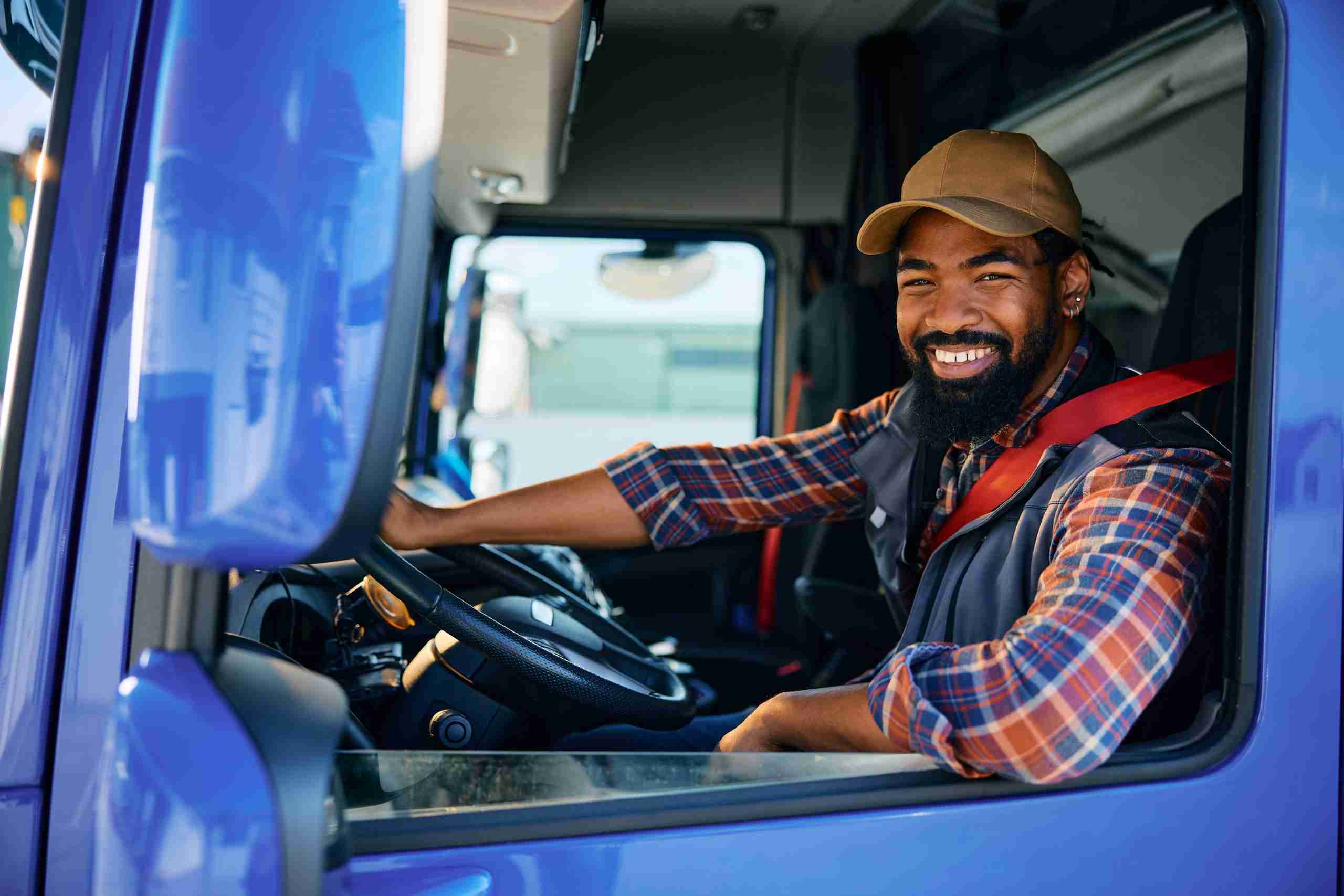 A man in a plaid shirt and cap sits in the driver's seat of a truck, smiling at the camera.