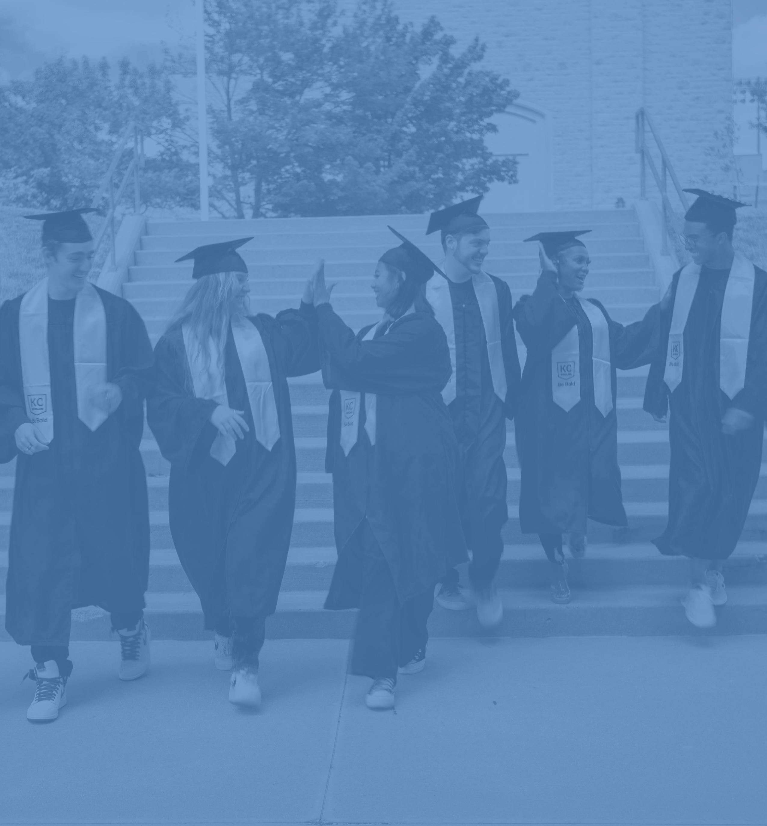 Five graduates wearing caps and gowns walk joyfully down a set of stairs, celebrating the start of their new careers. Some are giving high-fives while others are conversing and smiling.