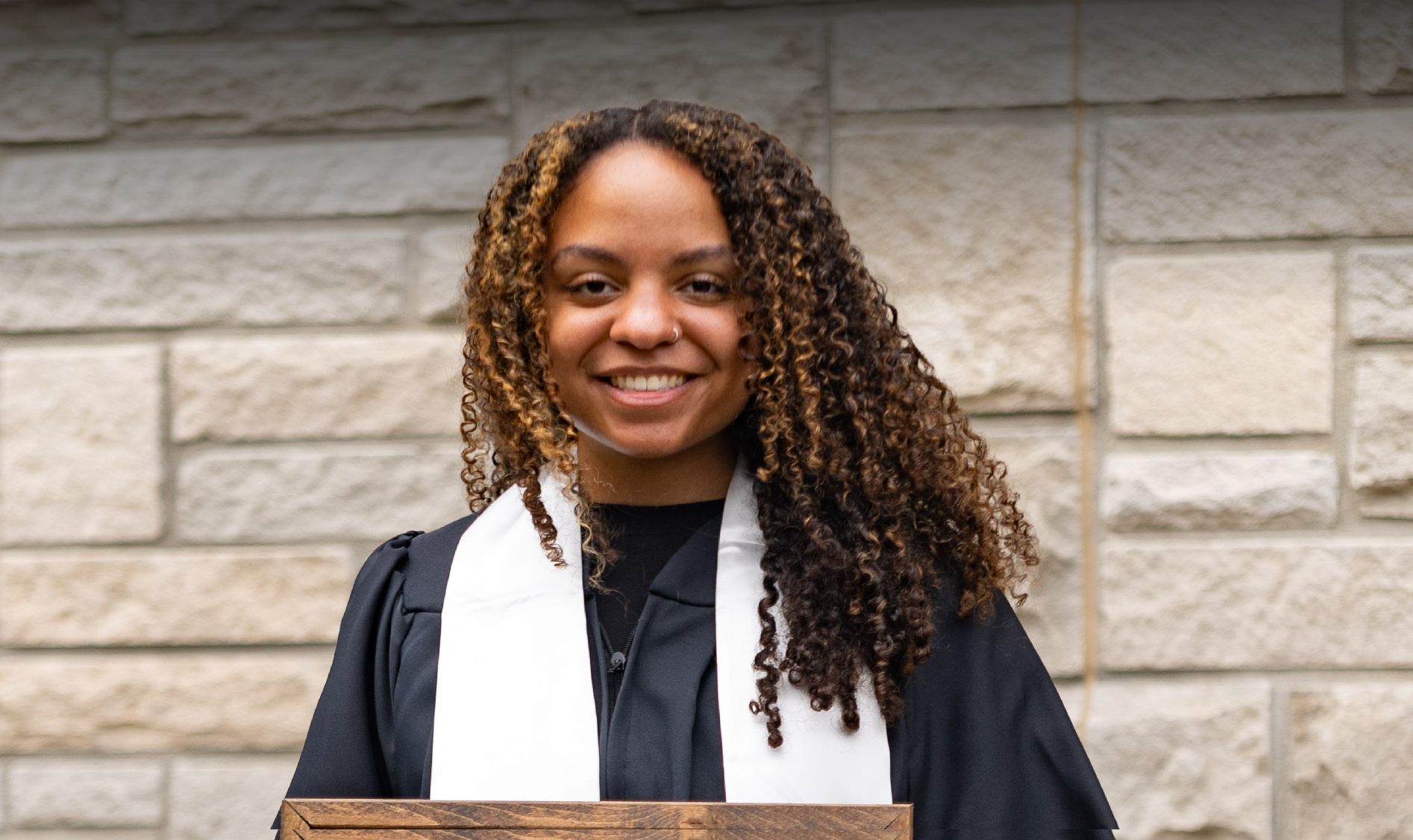 A person with curly hair, wearing a black graduation gown and white stole, stands in front of a stone wall holding a wooden plaque, celebrating the achievements that have paved the way for new job opportunities.