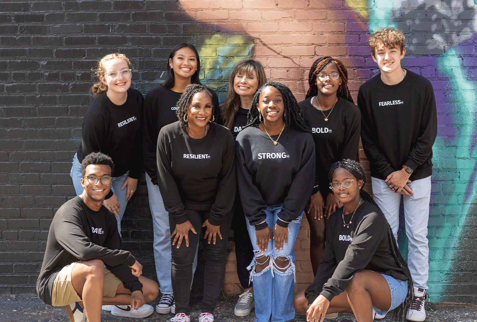 A group of nine young people, dressed in black shirts with various motivational words, stands and kneels in front of a colorful mural on a brick wall, smiling at the camera. Their bright smiles reflect the excitement for their future careers made possible through scholarships.