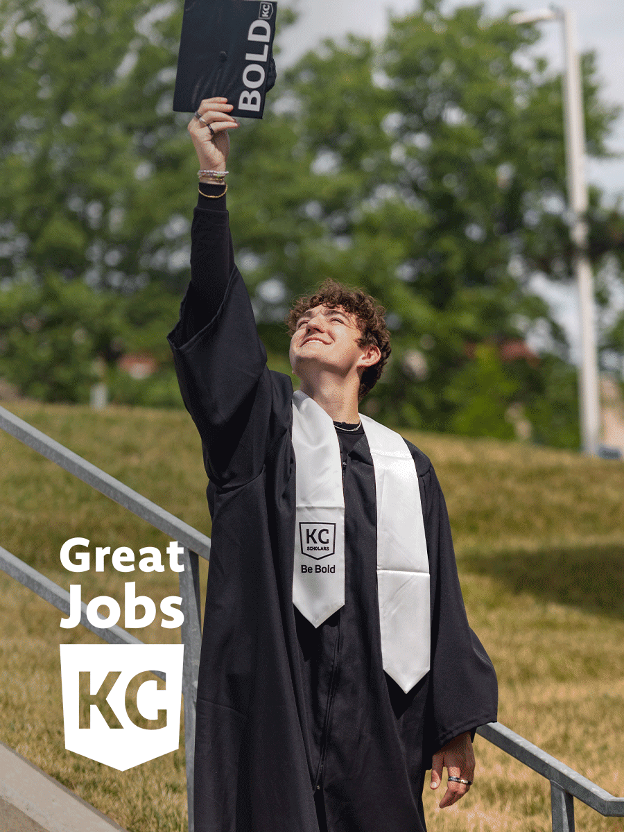 A young graduate in a black gown and white stole holds up a graduation cap, standing outdoors on steps with a "Great Jobs KC" sign next to him, ready to embark on his career.