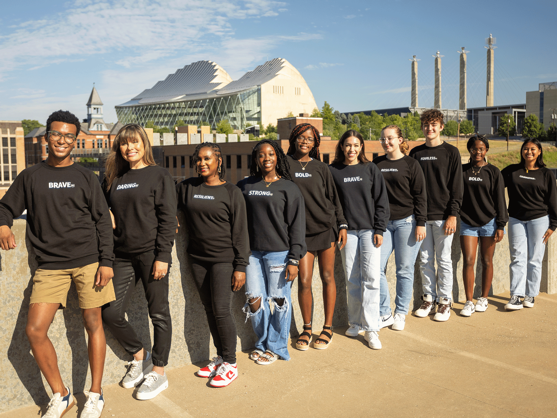 A diverse group of ten young adults stand in a line outdoors, all wearing black sweatshirts with empowering words. A modern building and three tall smokestacks are in the background under a clear sky, symbolizing their aspirations for careers as they pursue scholarships and job opportunities.