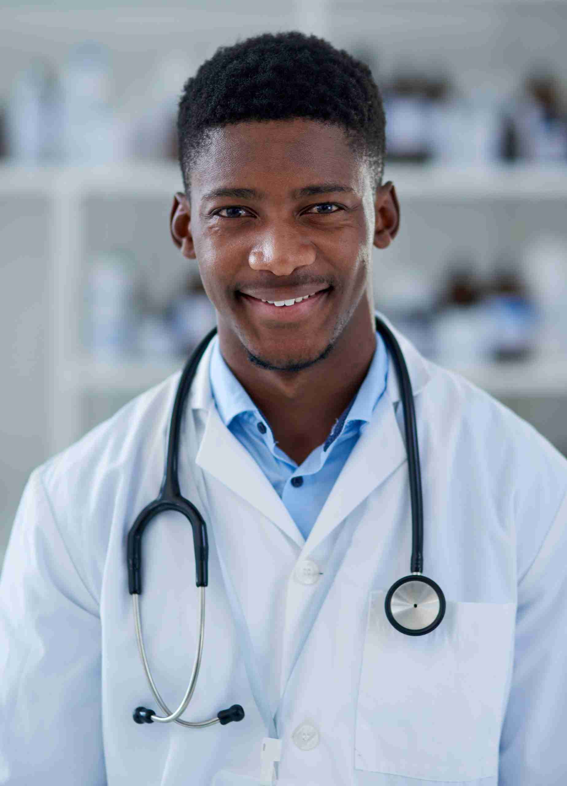 A person wearing a white lab coat and stethoscope, standing in a medical or laboratory setting with shelves in the background, perhaps contemplating available scholarships or considering job opportunities in the healthcare field.