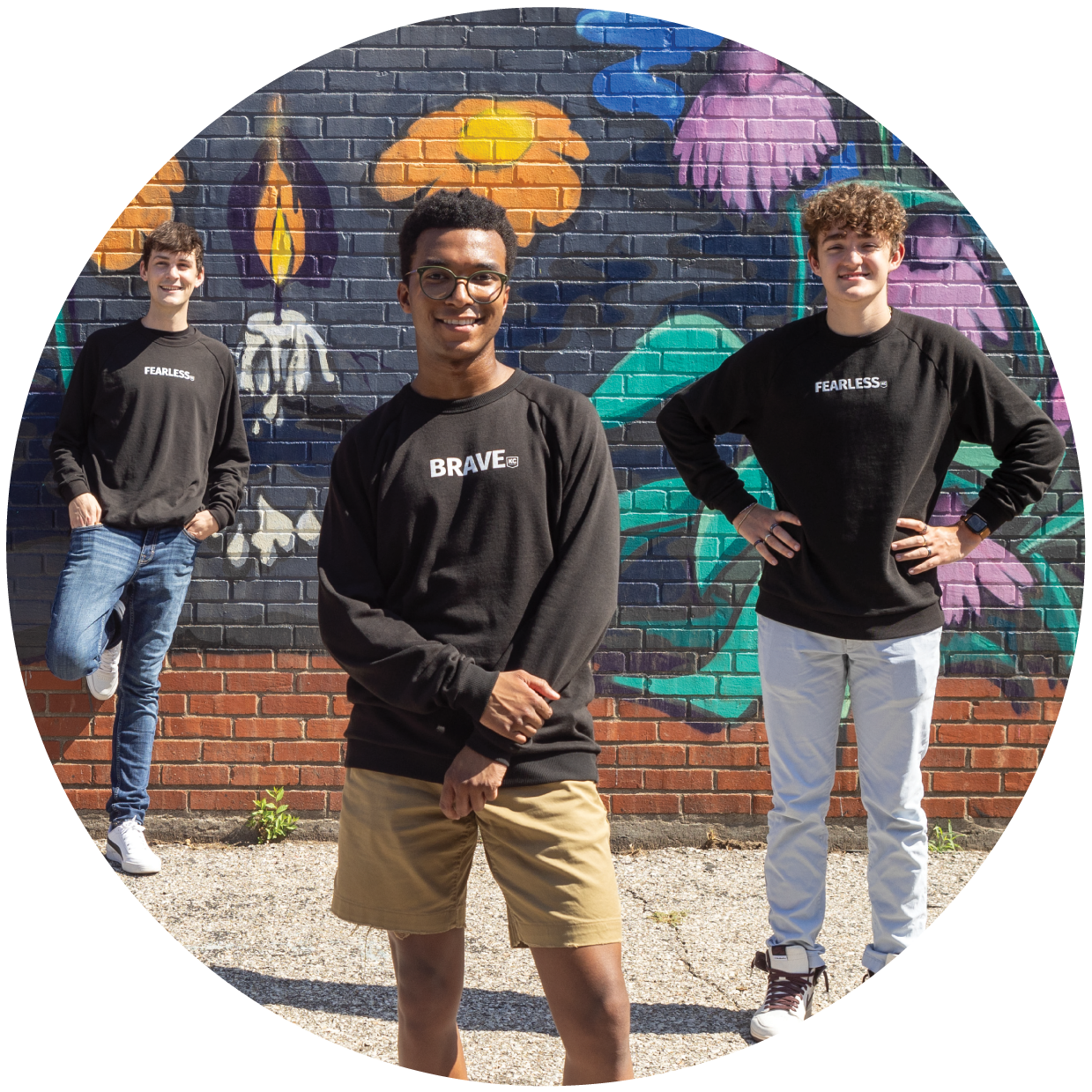 Three young men pose in front of a colorful mural on a brick wall. The man in the center wears glasses and a "BRAVE" shirt, while the others wear "FEARLESS" shirts, united by their pursuit of scholarships to advance their careers.