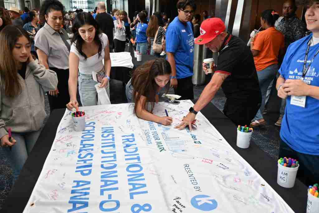 A diverse group of people signs a large banner titled "Traditional & Institutional Scholarship Awards" at an event. The table has numerous markers, and event staff in blue shirts can be seen in the background, highlighting opportunities for scholarships that pave the way for future careers.