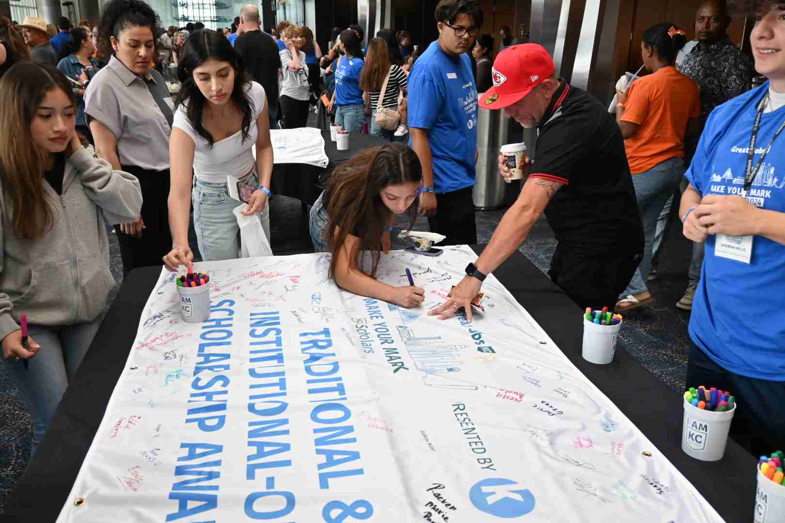 A diverse group of people signs a large banner titled "Traditional & Institutional Scholarship Awards" at an event. The table has numerous markers, and event staff in blue shirts can be seen in the background, highlighting opportunities for scholarships that pave the way for future careers.