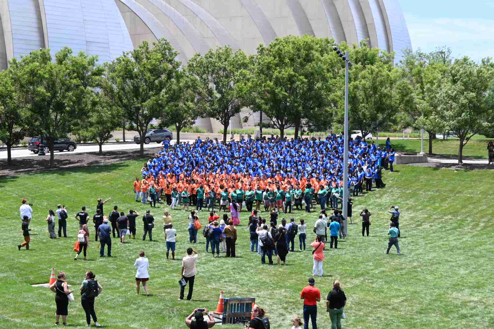 A large group of people in blue, green, and orange shirts are gathered on a grassy field, forming a formation. Onlookers and photographers surround them. Trees and a modern building are in the background, highlighting the event which celebrates scholarship achievements and promising career paths.