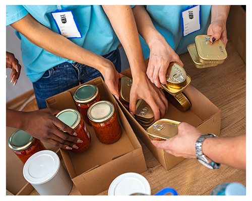 Volunteers pack canned goods and jars of food into boxes. Multiple hands are seen working together.