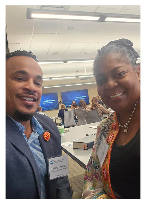 Two people smile for a selfie in a conference room. Both wear name tags, and a presentation is ongoing in the background.