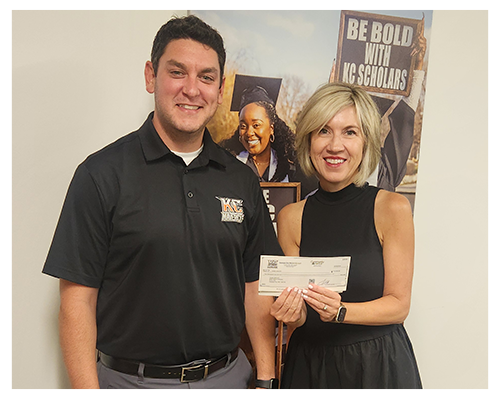 Two people stand together, smiling, in front of a poster; one holds a check. The poster reads "BE BOLD WITH KC SCHOLARS.