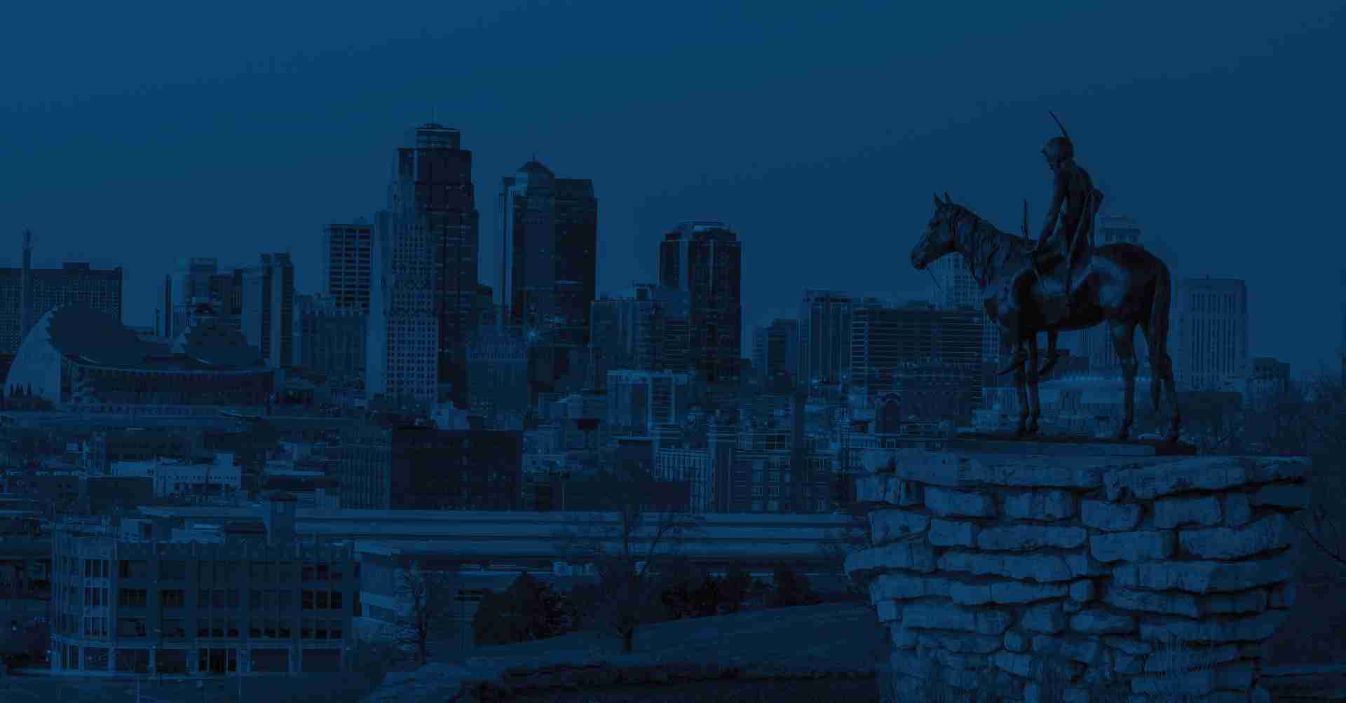A statue of a person riding a horse stands on a rocky pedestal with a city skyline in the background under a dark blue sky.