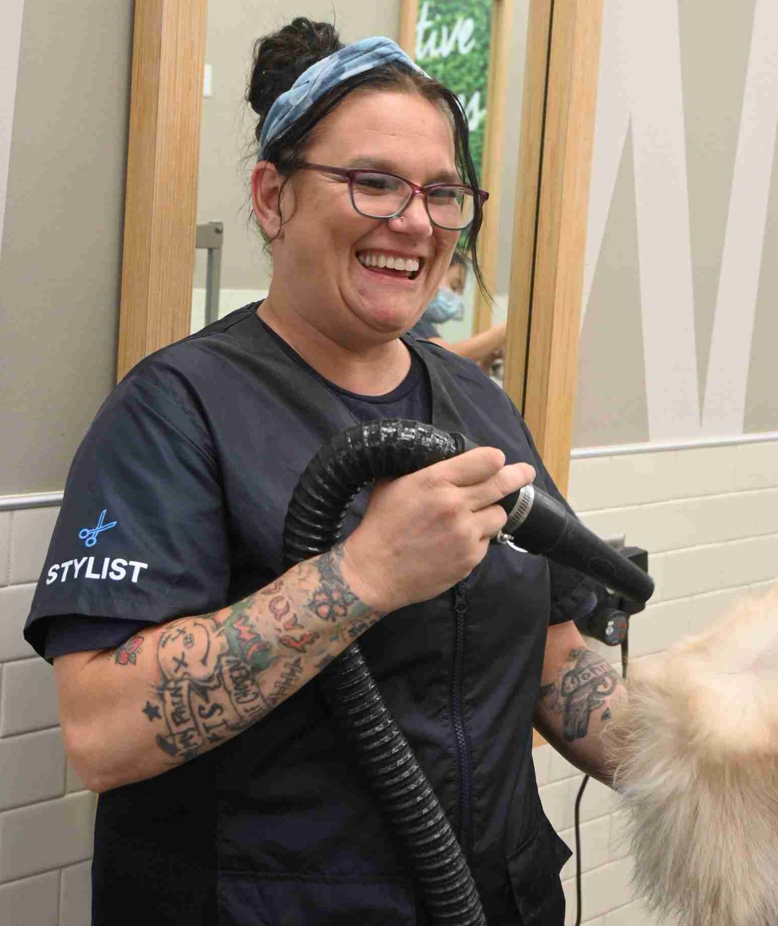 Person with tattoos and glasses, wearing a stylist uniform, smiles while holding a grooming tool near a dog's fur in a salon setting.