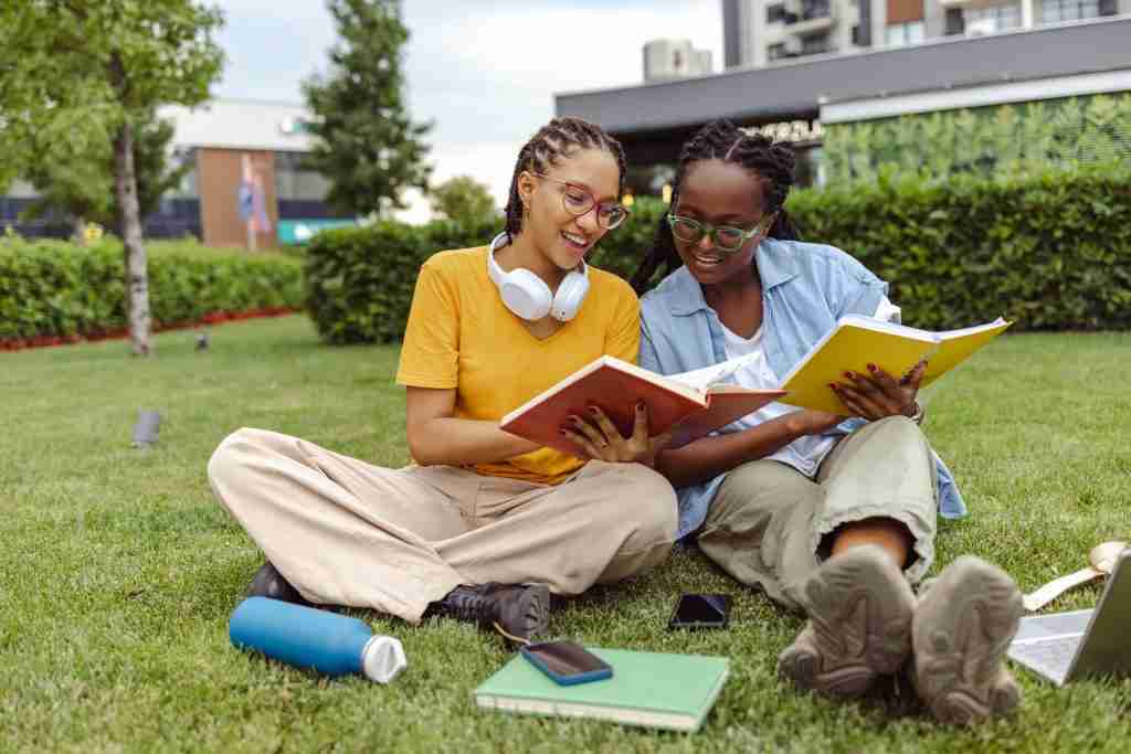 Two people sit on grass, smiling and reading books together. A phone, headphones, and water bottle lie nearby. Trees and a building are in the background.