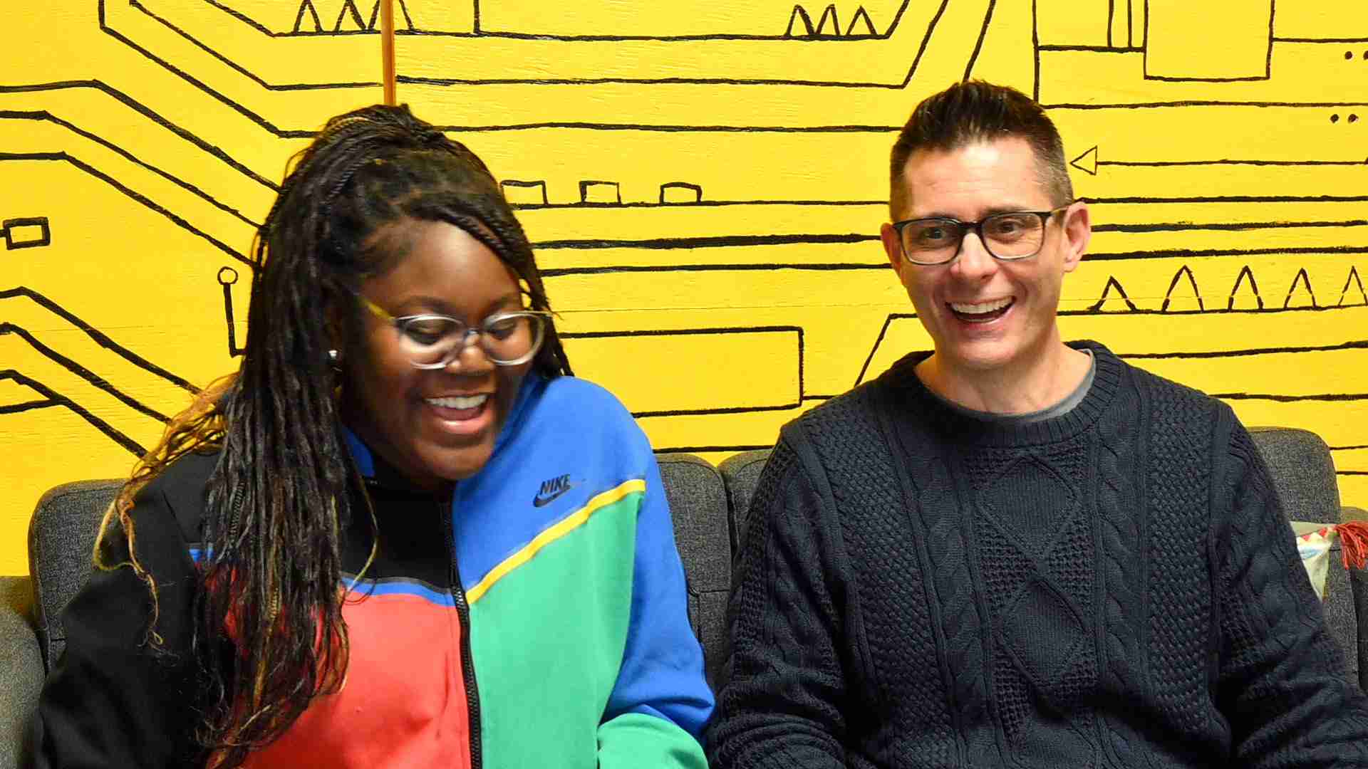 Two adult learners sit on a couch in front of a yellow wall with black geometric designs, both smiling and wearing glasses.