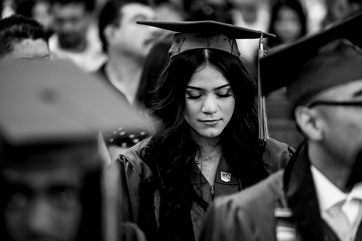 A young woman in a graduation cap and gown looks down, surrounded by fellow graduates at a ceremony. This black and white image captures the spirit highlighted in the Kansas City education impact newsletter.