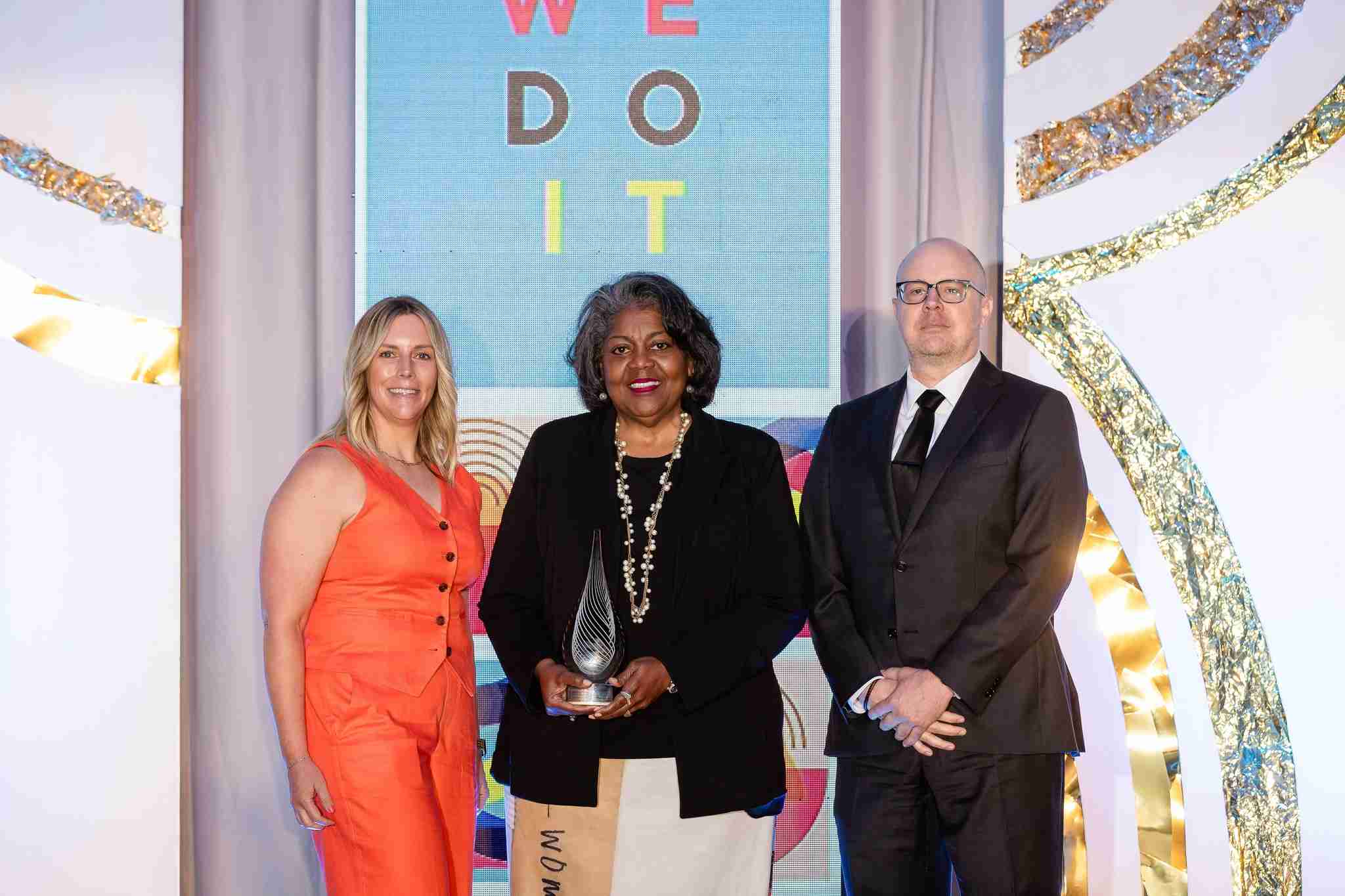 Three people stand posing at an awards event; the woman in the center holds a glass trophy, with a "WE DO IT" sign and mention of the Kansas City education impact newsletter in the background.