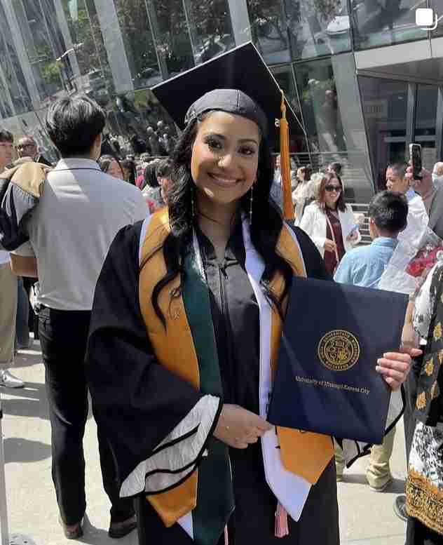 A graduate in cap and gown holds a diploma folder while smiling at an outdoor graduation ceremony, surrounded by people.