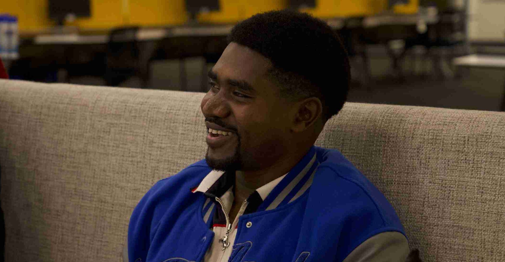 A man wearing a blue jacket sits on a beige couch and smiles in a modern office setting with desks and yellow dividers, appearing relaxed and free from financial stress.
