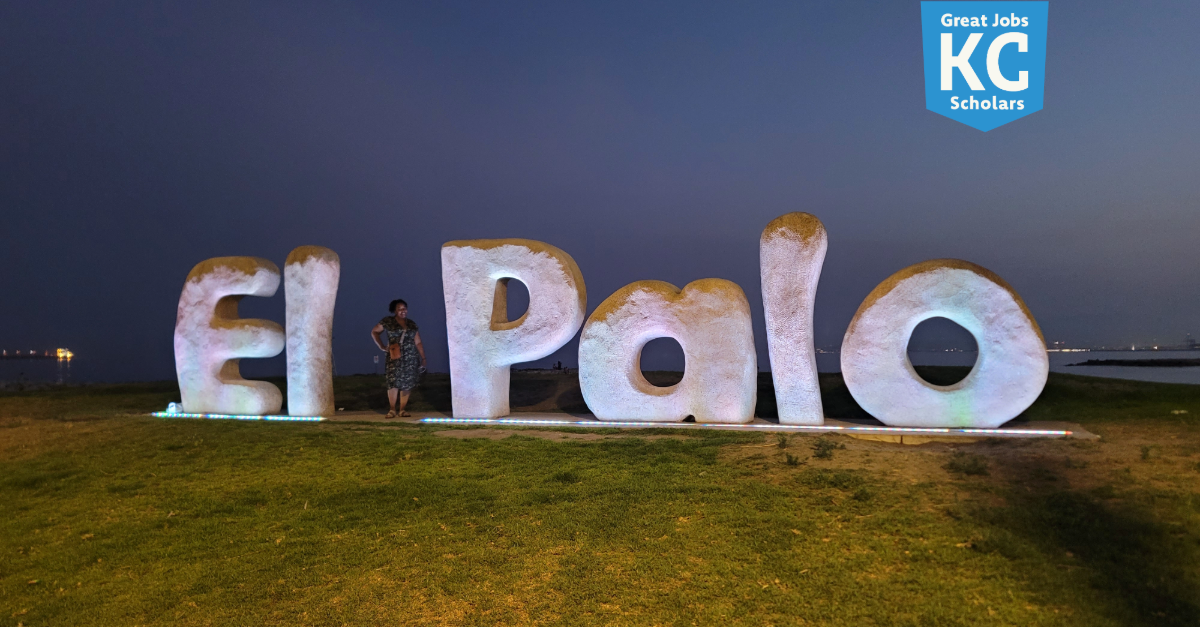 Large illuminated "El Palo" sign on grass at dusk near water, with a person standing between the letters; KC Scholars logo in top right corner, symbolizing hope and relief from financial stress.