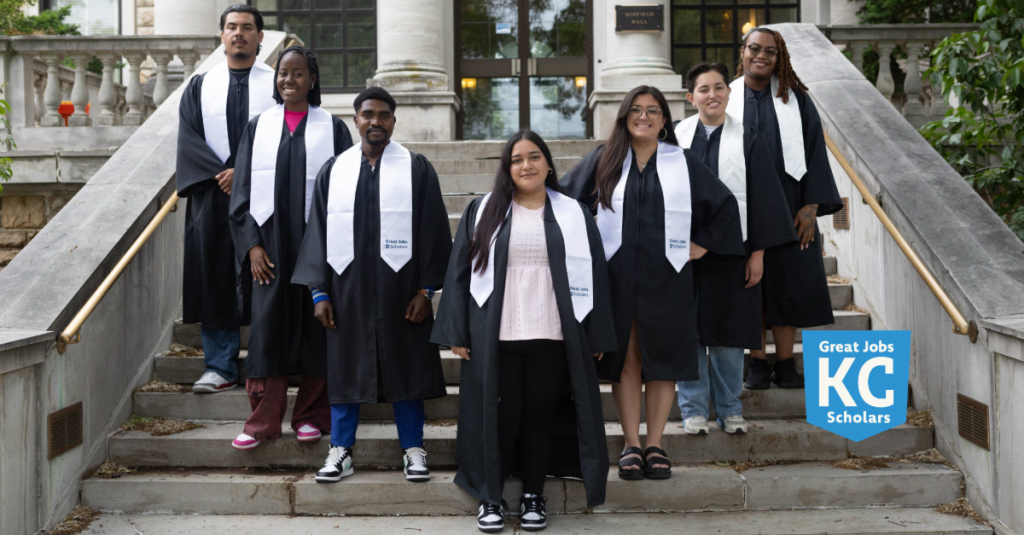Seven graduates in caps and gowns stand on outdoor steps, proudly wearing white stoles that read “Great Jobs KC Scholars”—a celebration of success achieved despite financial stress. A blue sign with the program’s logo appears in the lower right corner.