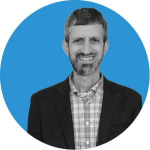 Man with short dark hair and beard, wearing a checked shirt and dark blazer, smiling in front of a blue circular background—perfect for an annual report profile.