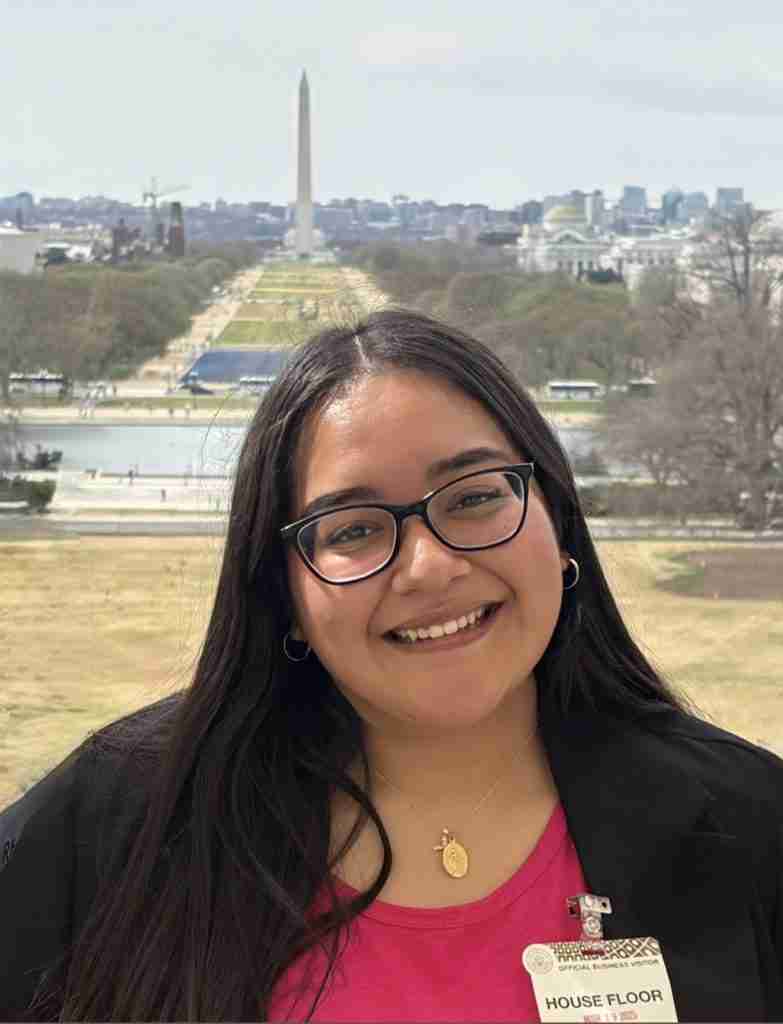 A woman with long dark hair and glasses smiles at the camera, embodying resilience and reflection, with the Washington Monument and National Mall in the background.