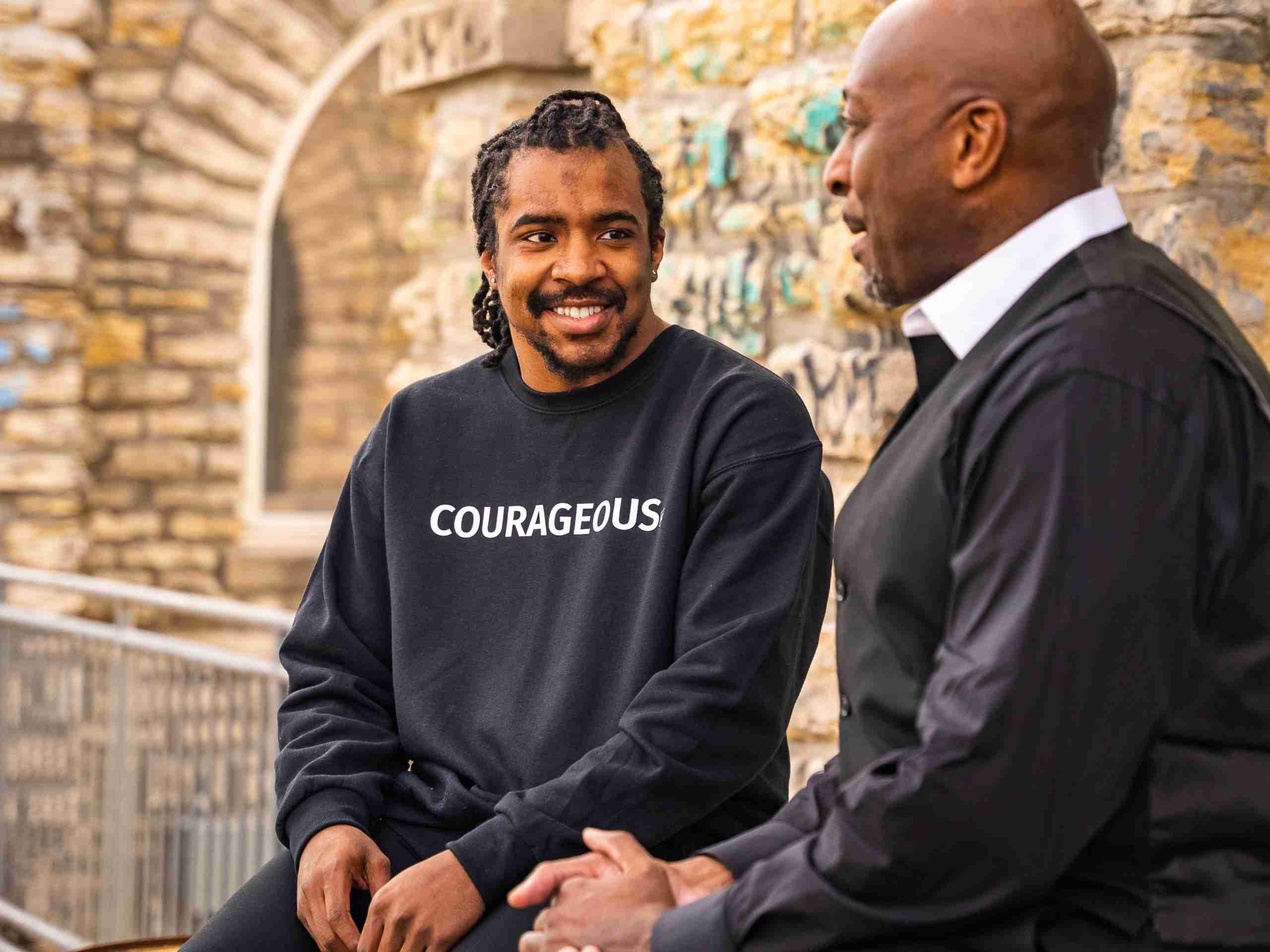Two men sit and talk near a stone wall; one wears a black sweatshirt with the word "COURAGEOUS" printed on it and smiles at the other man, who wears a black vest and shirt.