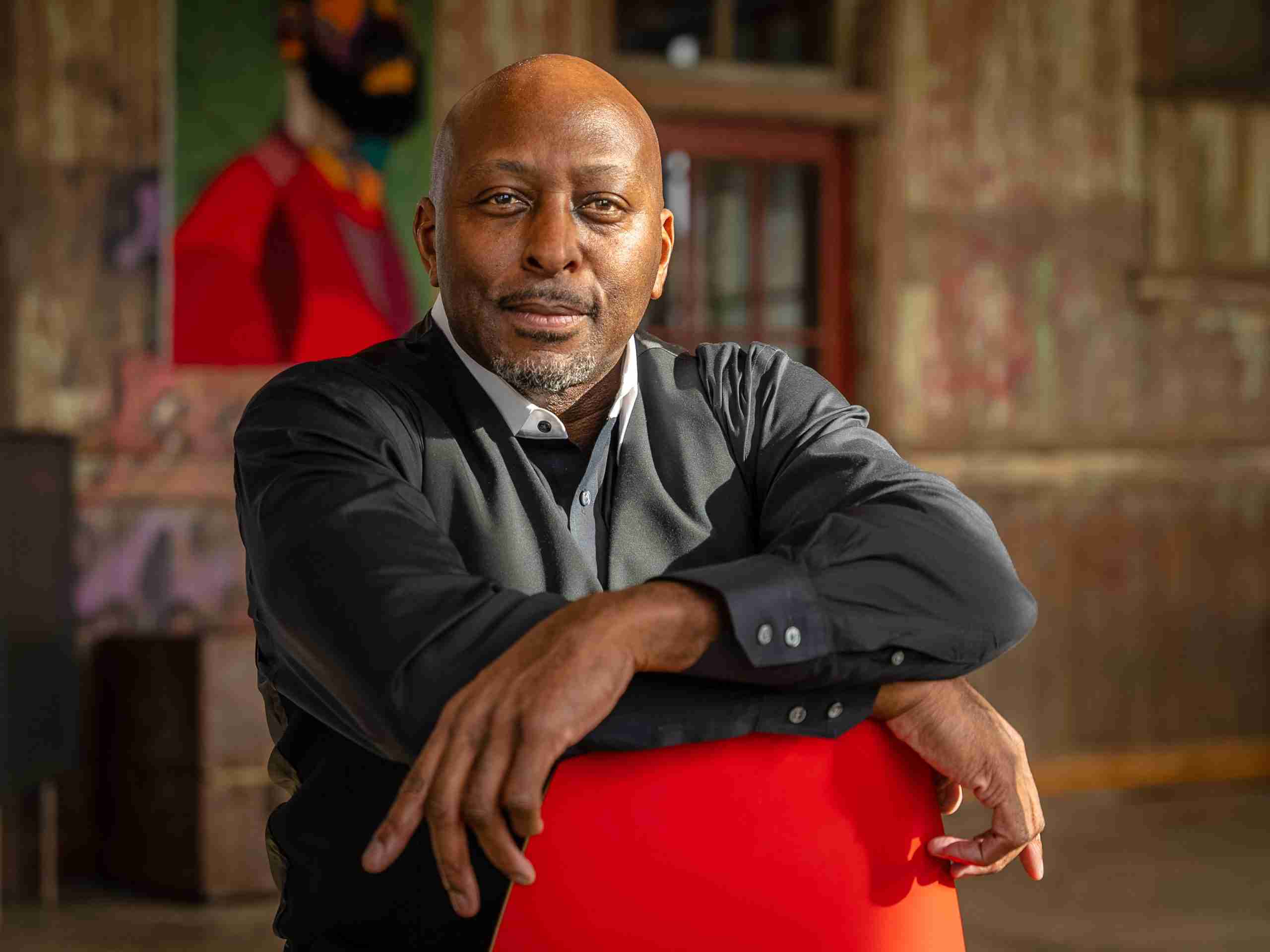 A man in a black shirt sits with his arms resting on the back of a red chair in a rustic room with a painting on the wall behind him.
