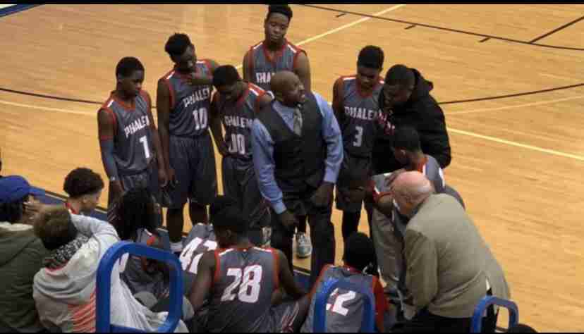 A high school basketball team in gray uniforms gathers around their coach during a timeout on a gym court, with spectators seated nearby.