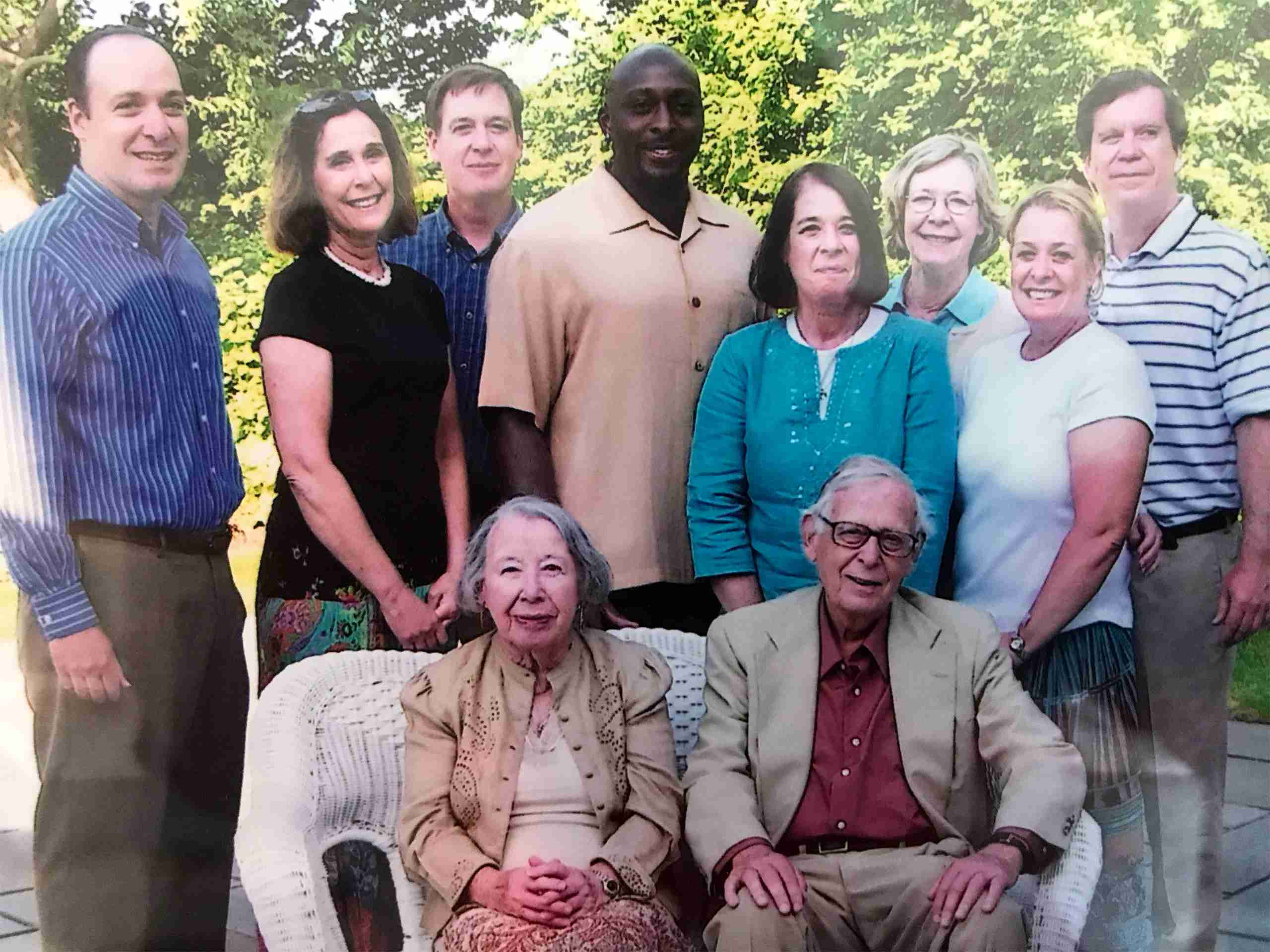 A group of eight adults stands behind an elderly man and woman seated on a wicker bench outdoors with green trees in the background.